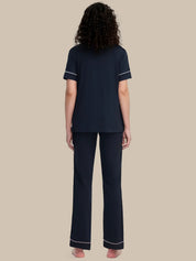 A woman viewed from the back wearing a navy blue short-sleeve pajama top and matching wide-leg pajama pants with thin white piping trim on the sleeve cuffs and hems, set against a solid beige background.