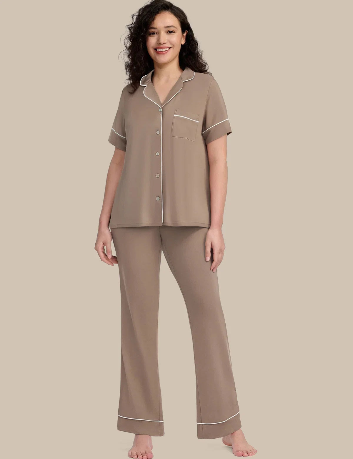 A woman modeling a two-piece pajama set in taupe with white piping details, consisting of a short-sleeve button-up top and matching wide-leg pants, set against a solid beige background.