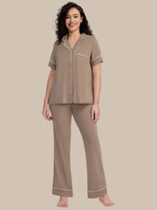 A woman modeling a two-piece pajama set in taupe with white piping details, consisting of a short-sleeve button-up top and matching wide-leg pants, set against a solid beige background.
