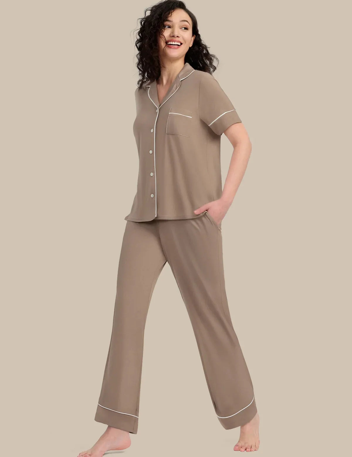 A woman modeling a two-piece pajama set in taupe with white piping details, consisting of a short-sleeve button-up top and matching wide-leg pants, set against a solid beige background.