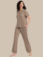 A woman modeling a two-piece pajama set in taupe with white piping details, consisting of a short-sleeve button-up top and matching wide-leg pants, set against a solid beige background.