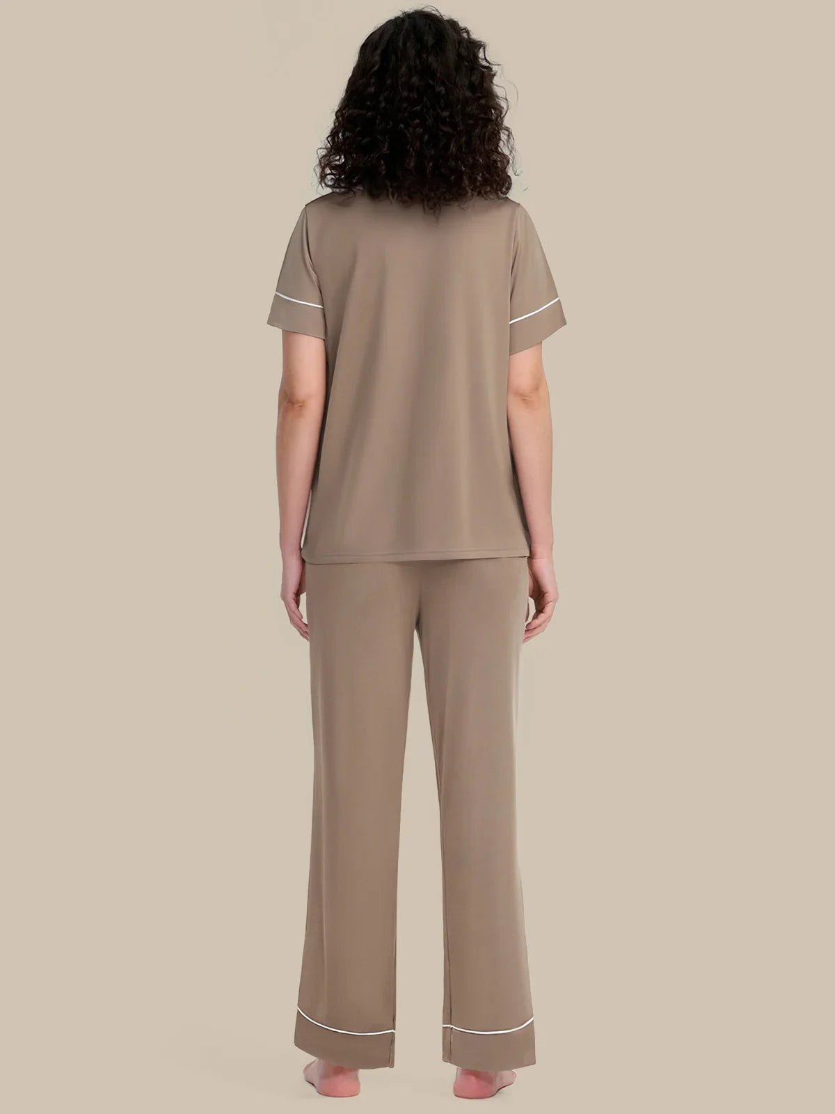 A woman viewed from the back wearing a matching taupe lounge set with white piping trim on the sleeves and pants cuffs, against a solid beige background.