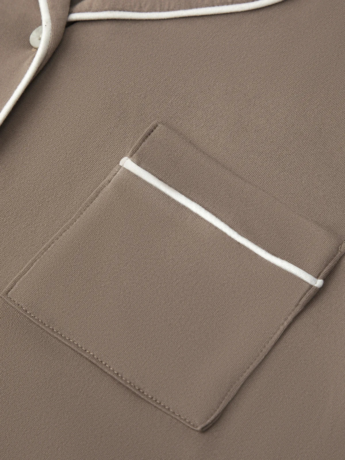 Close-up of a taupe brown garment featuring a chest pocket with white piping and a matching white button, showing the fabric texture.