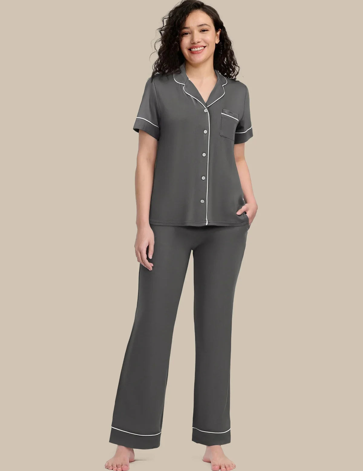 A woman modeling a two-piece gray pajama set with white piping details, consisting of a short-sleeve button-up top and matching pants, against a solid beige background.