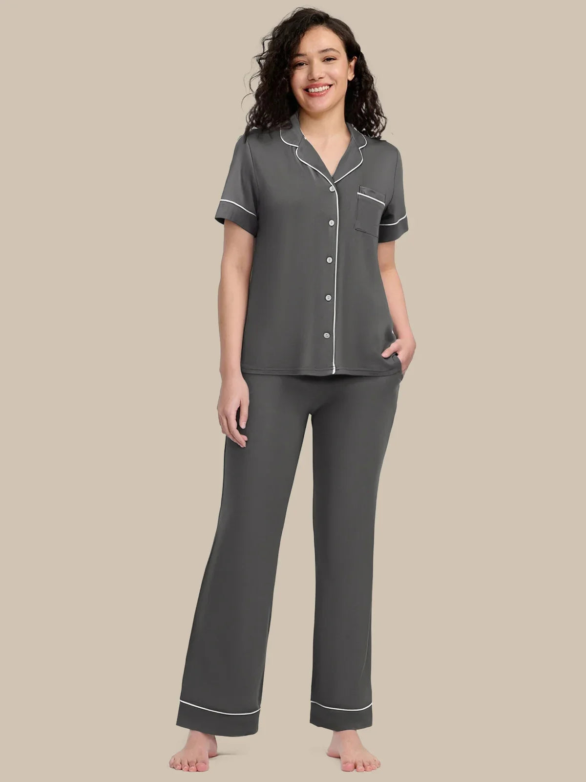 A woman modeling a two-piece gray pajama set with white piping details, consisting of a short-sleeve button-up top and matching pants, against a solid beige background.