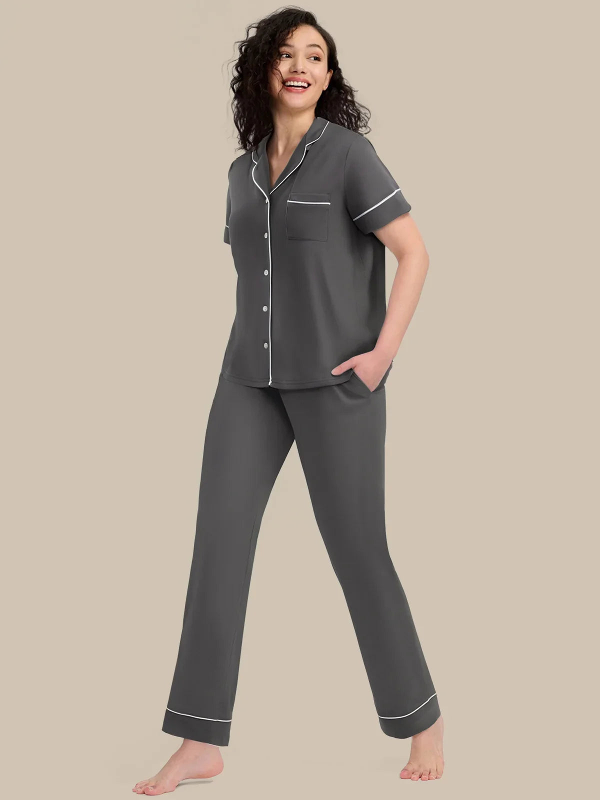 A woman modeling a dark gray short-sleeve pajama set with white piping details, consisting of a button-front top and matching pants, against a solid beige background.