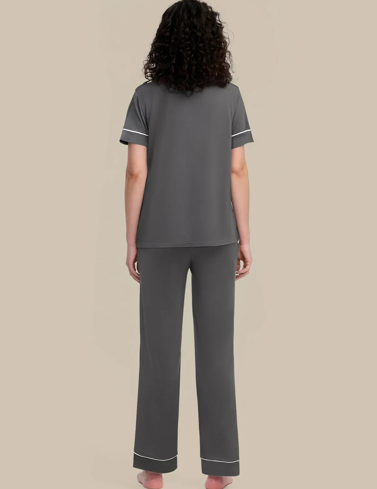 A woman viewed from the back wearing a dark gray two-piece pajama set with white piping trim on the sleeves and pants cuffs, against a solid beige background.