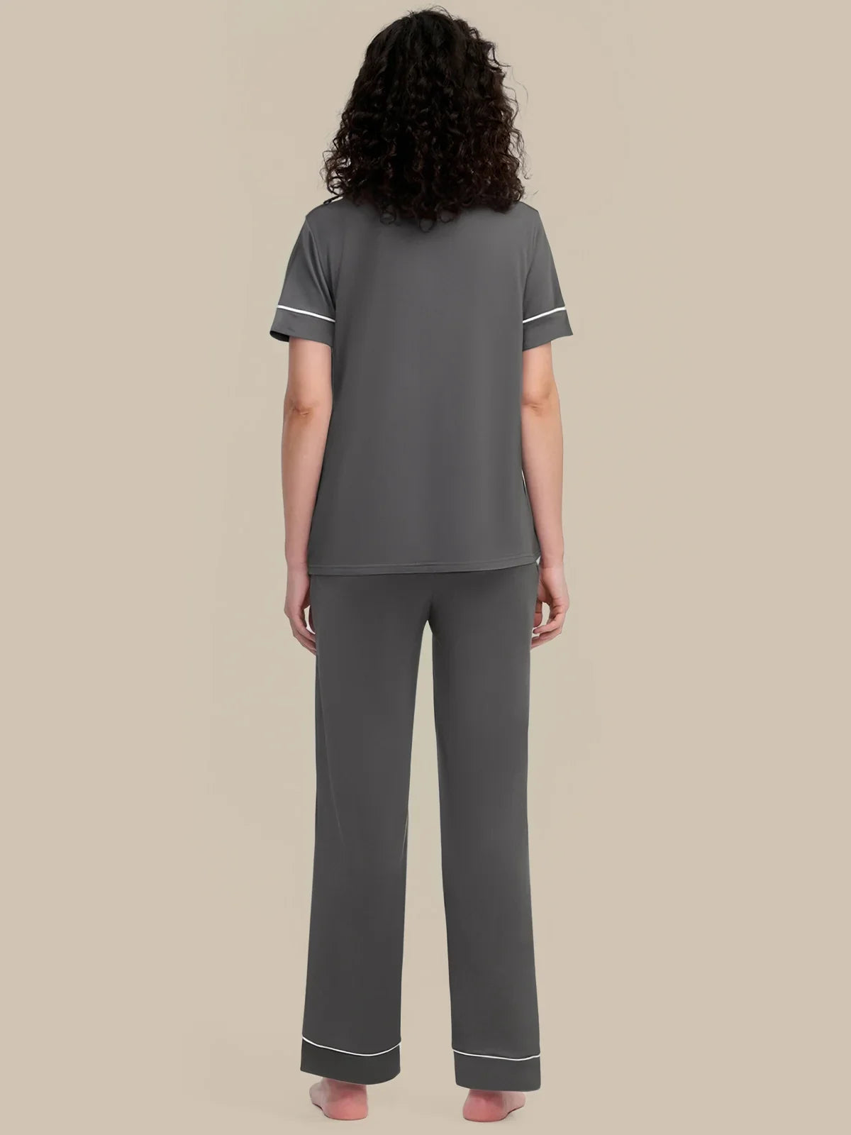 A woman viewed from the back wearing a dark gray two-piece pajama set with white piping trim on the sleeves and pants cuffs, against a solid beige background.