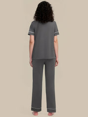 A woman viewed from the back wearing a dark gray two-piece pajama set with white piping trim on the sleeves and pants cuffs, against a solid beige background.