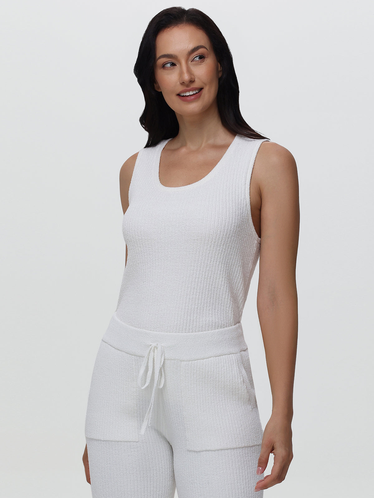 Woman wearing a white ribbed tank top and pants on a plain background