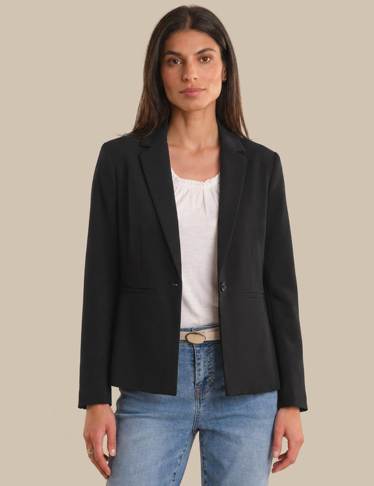 Woman wearing a black blazer over a white top and blue jeans on a beige background