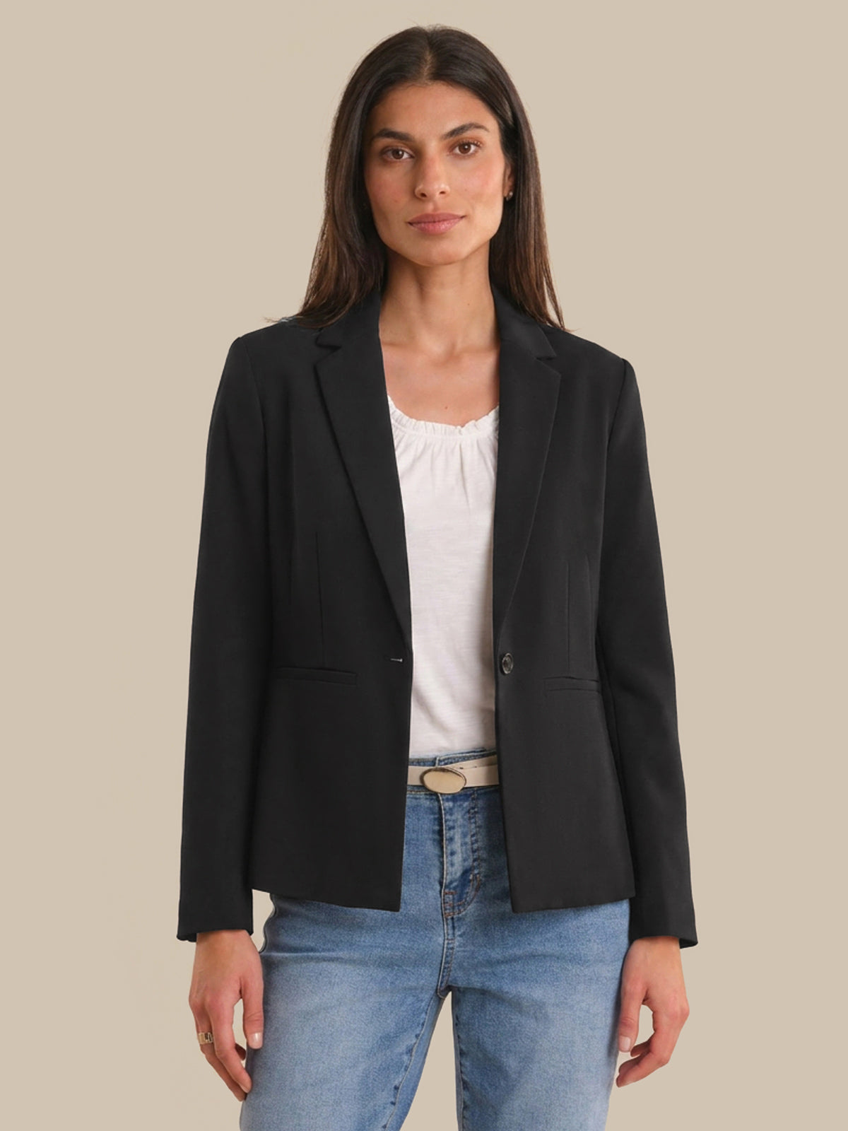 Woman wearing a black blazer over a white top and blue jeans on a beige background