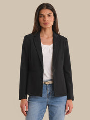 Woman wearing a black blazer over a white top and blue jeans on a beige background