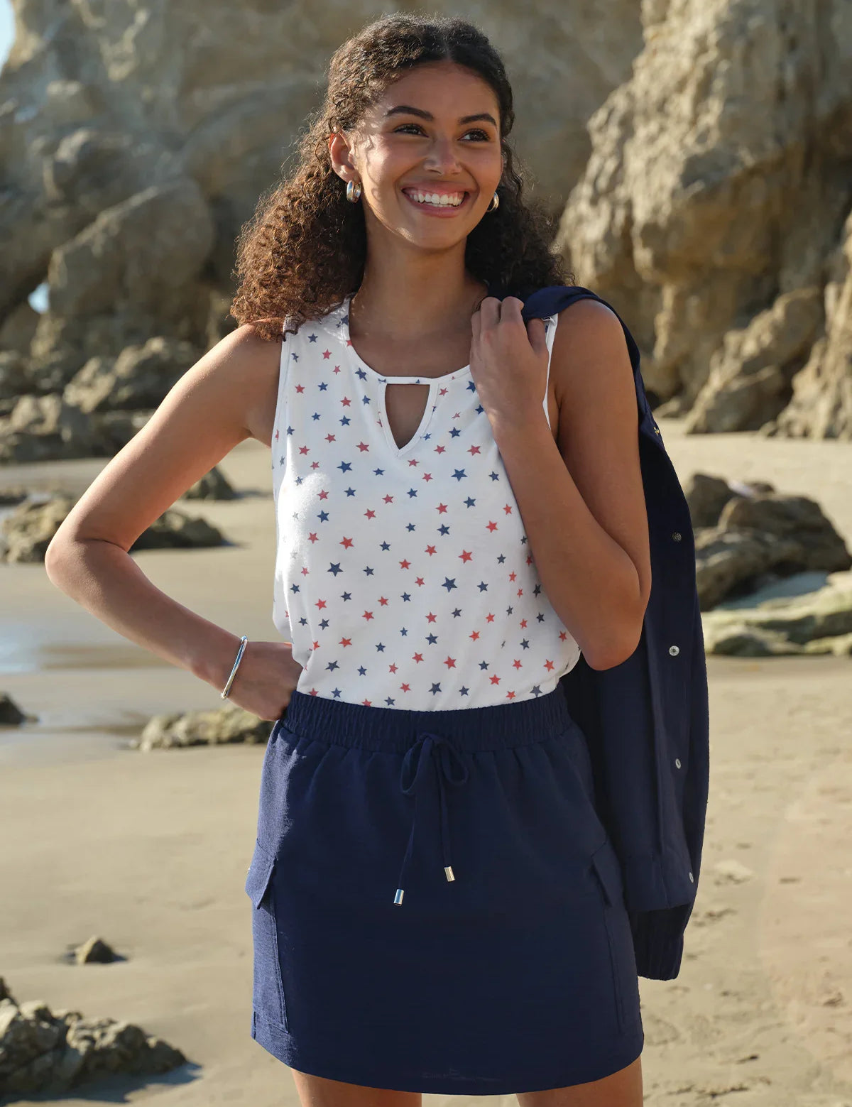 Woman standing on a beach wearing a white top with red and blue patterns and a navy skirt.