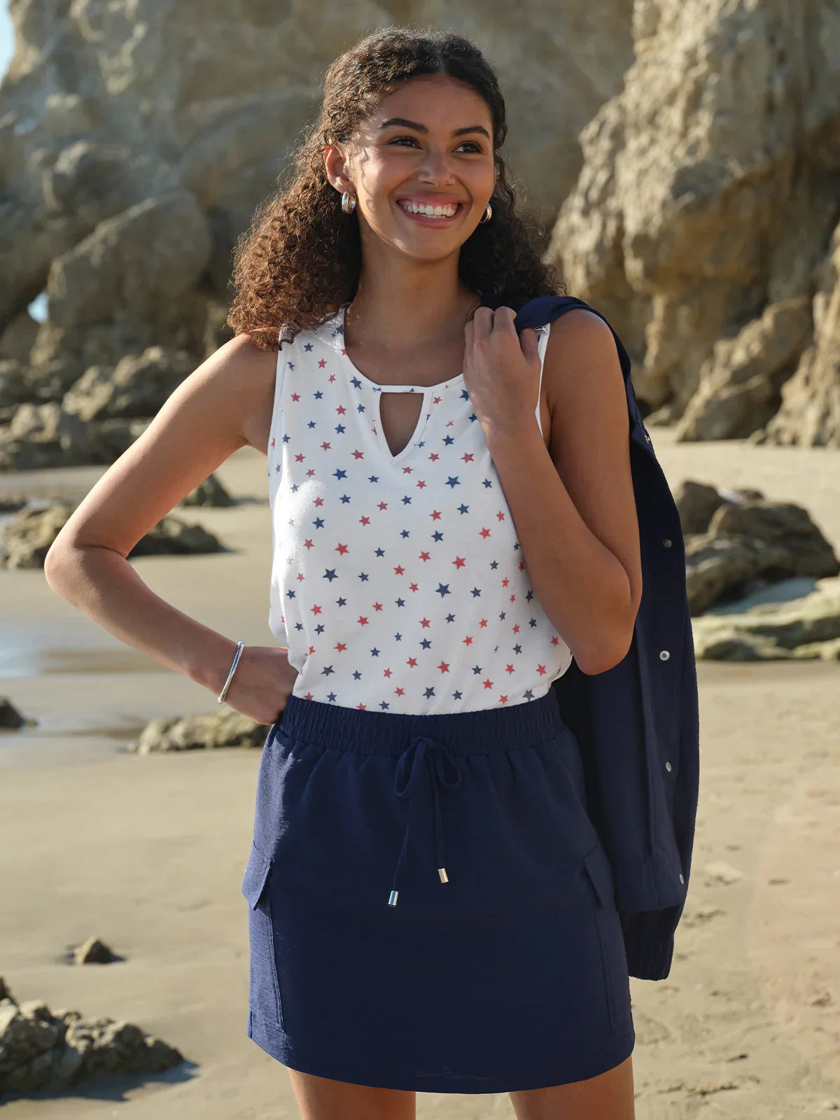 Woman standing on a beach wearing a white top with red and blue patterns and a navy skirt.