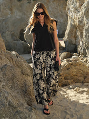 Woman in black sleeveless top and floral pants standing on a rocky beach.