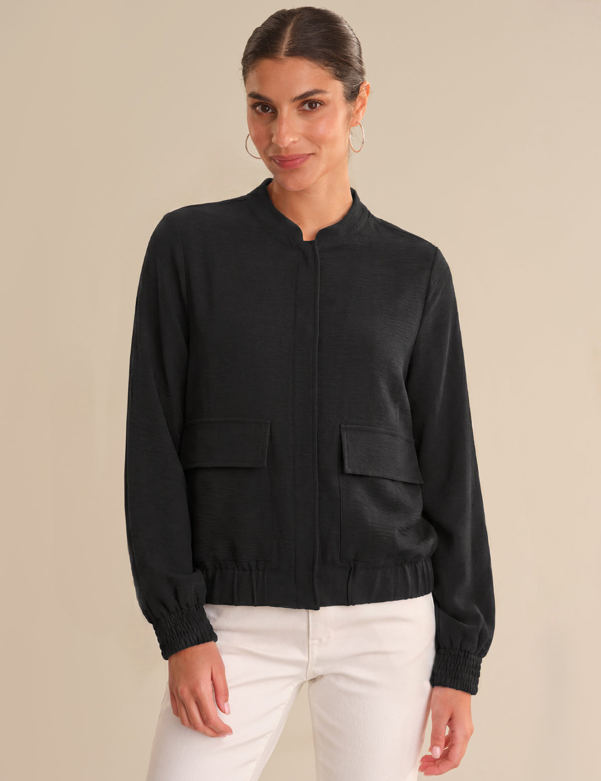 Woman wearing a black bomber jacket and white pants on a beige background