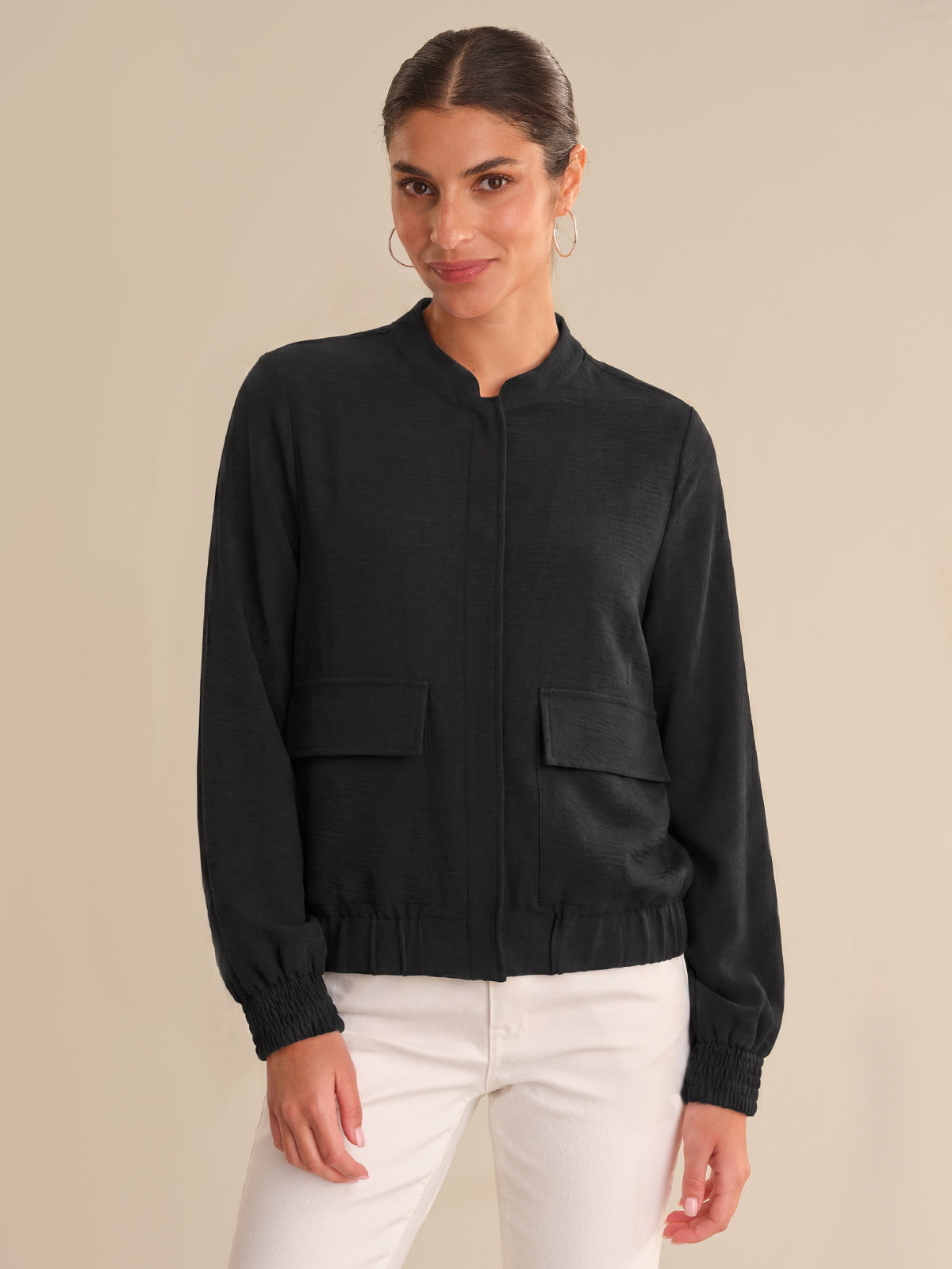 Woman wearing a black bomber jacket and white pants on a beige background