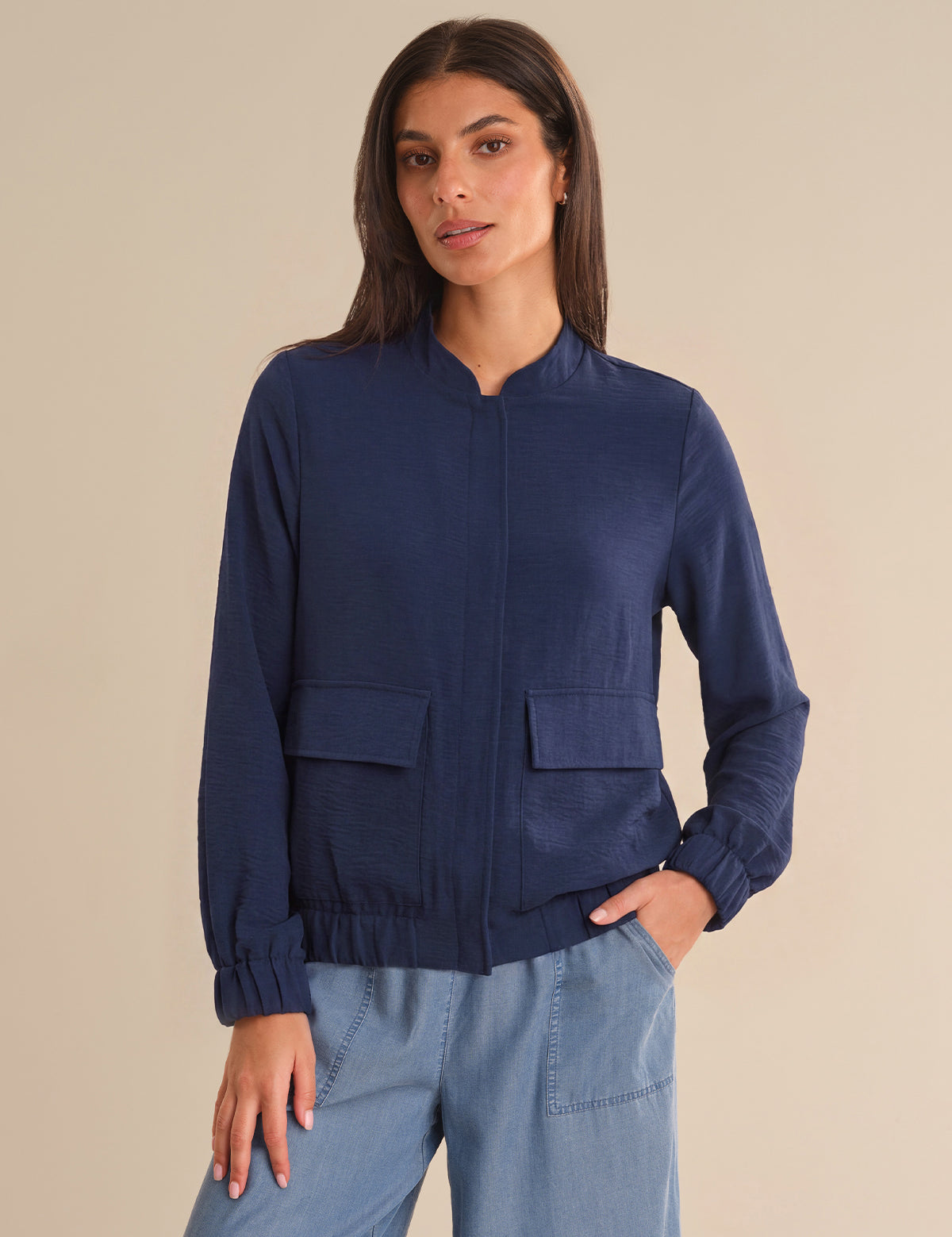 Woman wearing a navy blue bomber jacket and light blue pants against a beige background