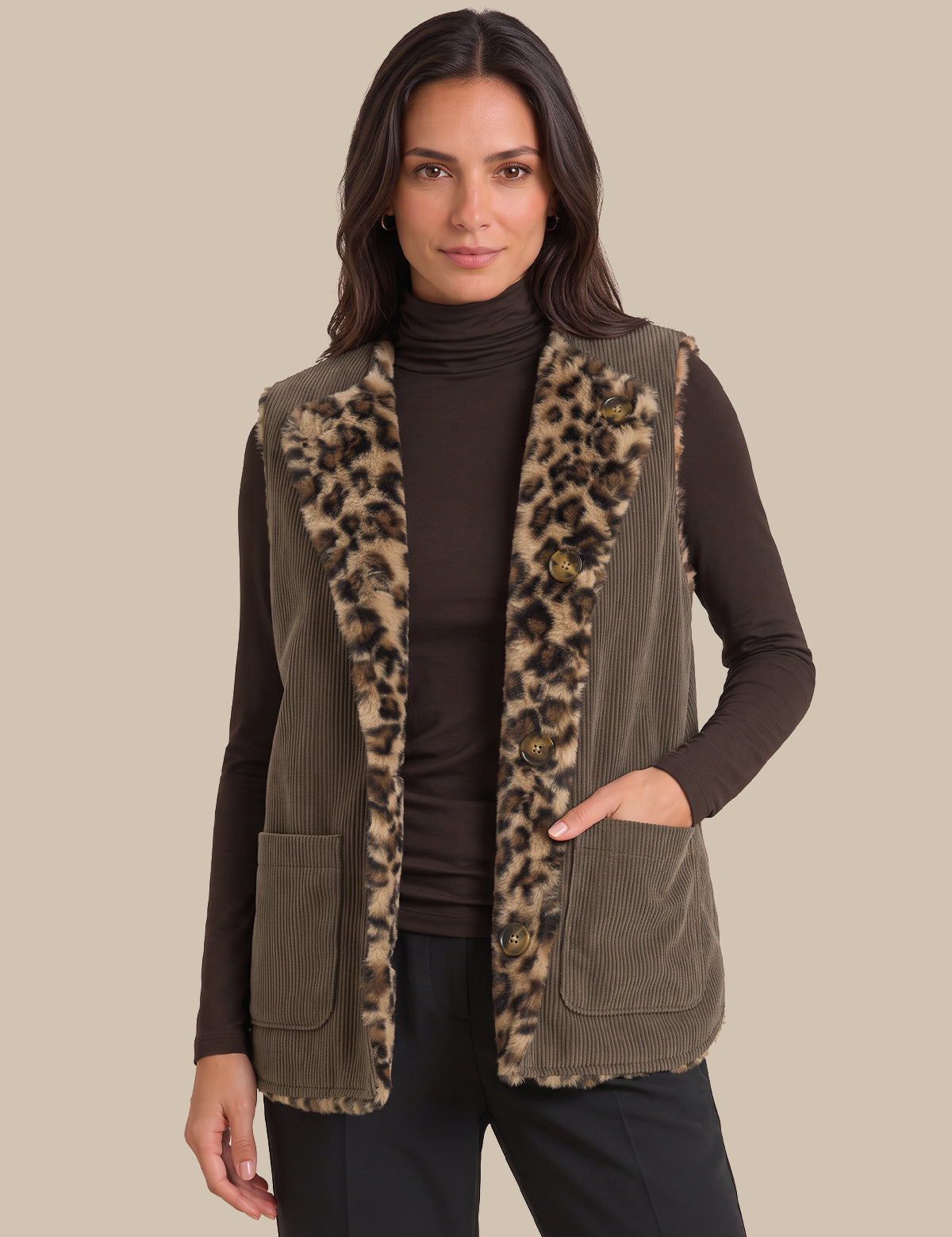 A woman wearing a brown corduroy vest with a leopard-print faux fur trim over a dark brown turtleneck sweater and black pants, set against a solid beige background.