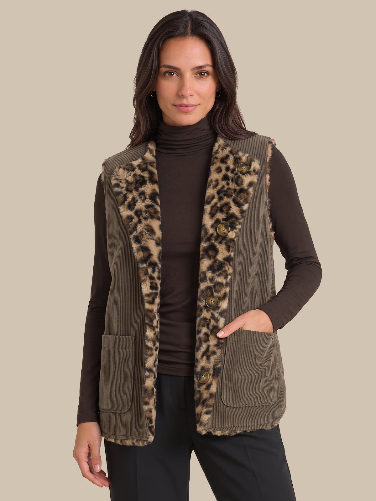 A woman wearing a brown corduroy vest with a leopard-print faux fur trim over a dark brown turtleneck sweater and black pants, set against a solid beige background.
