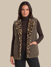 A woman wearing a brown corduroy vest with a leopard-print faux fur trim over a dark brown turtleneck sweater and black pants, set against a solid beige background.