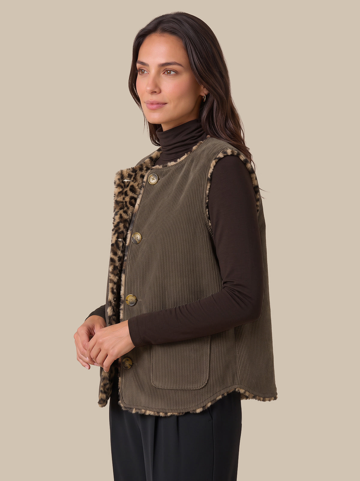 A woman modeling a corduroy vest with leopard-print trim over a dark brown turtleneck and black pants, set against a solid beige background.