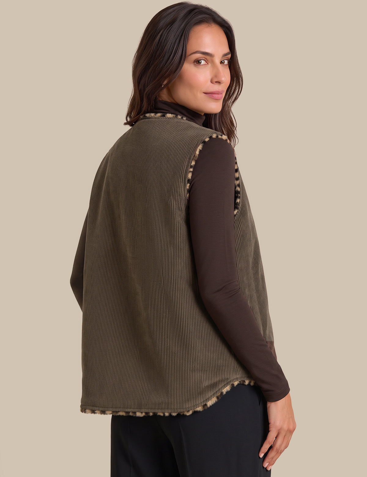 A woman shown from the back wearing an olive green ribbed sleeveless vest over a dark brown long-sleeve top and black pants, against a solid beige background.
