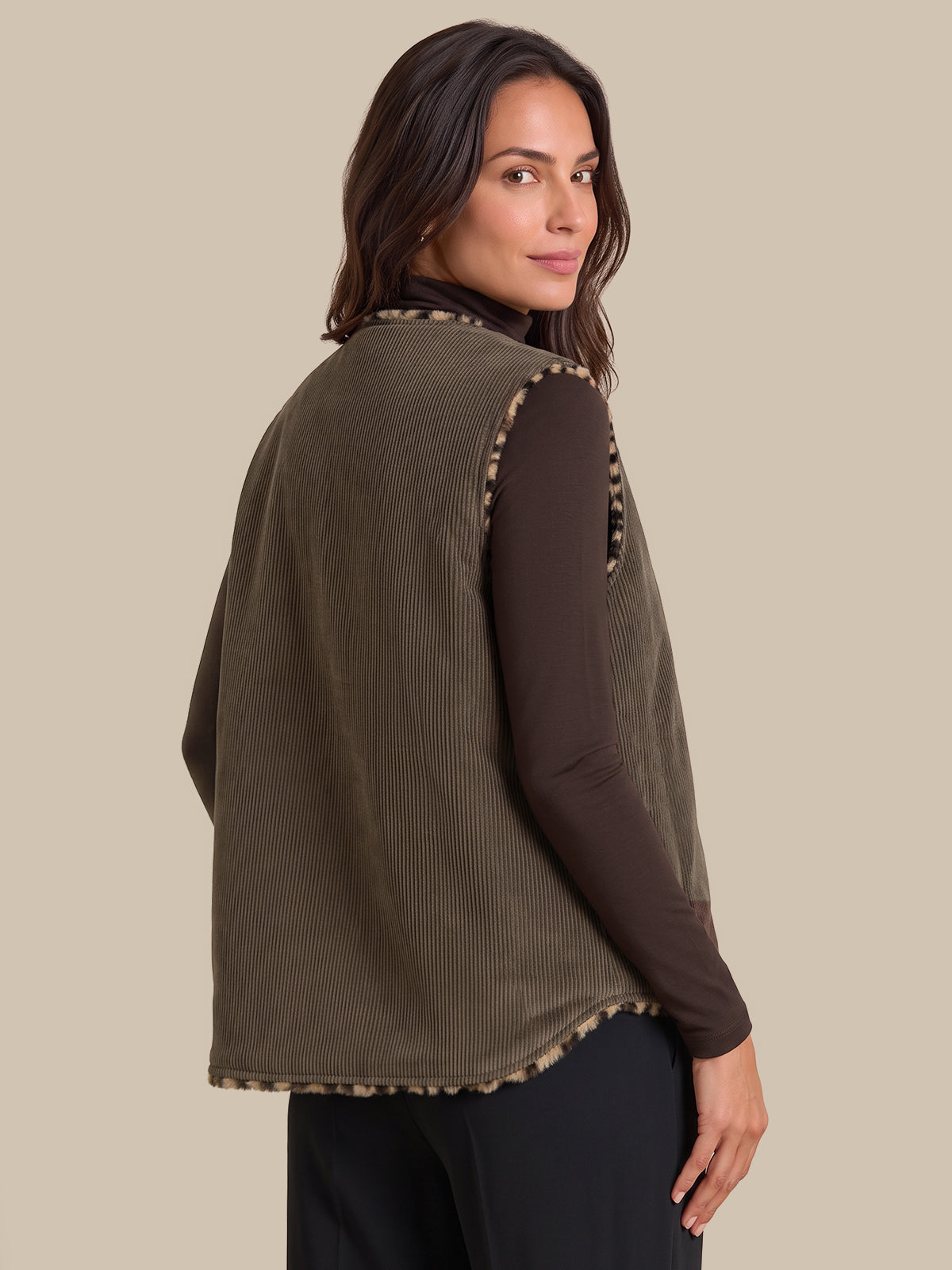 A woman shown from the back wearing an olive green ribbed sleeveless vest over a dark brown long-sleeve top and black pants, against a solid beige background.