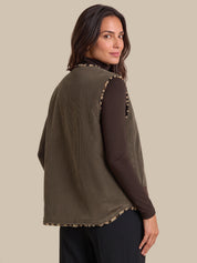 A woman shown from the back wearing an olive green ribbed sleeveless vest over a dark brown long-sleeve top and black pants, against a solid beige background.