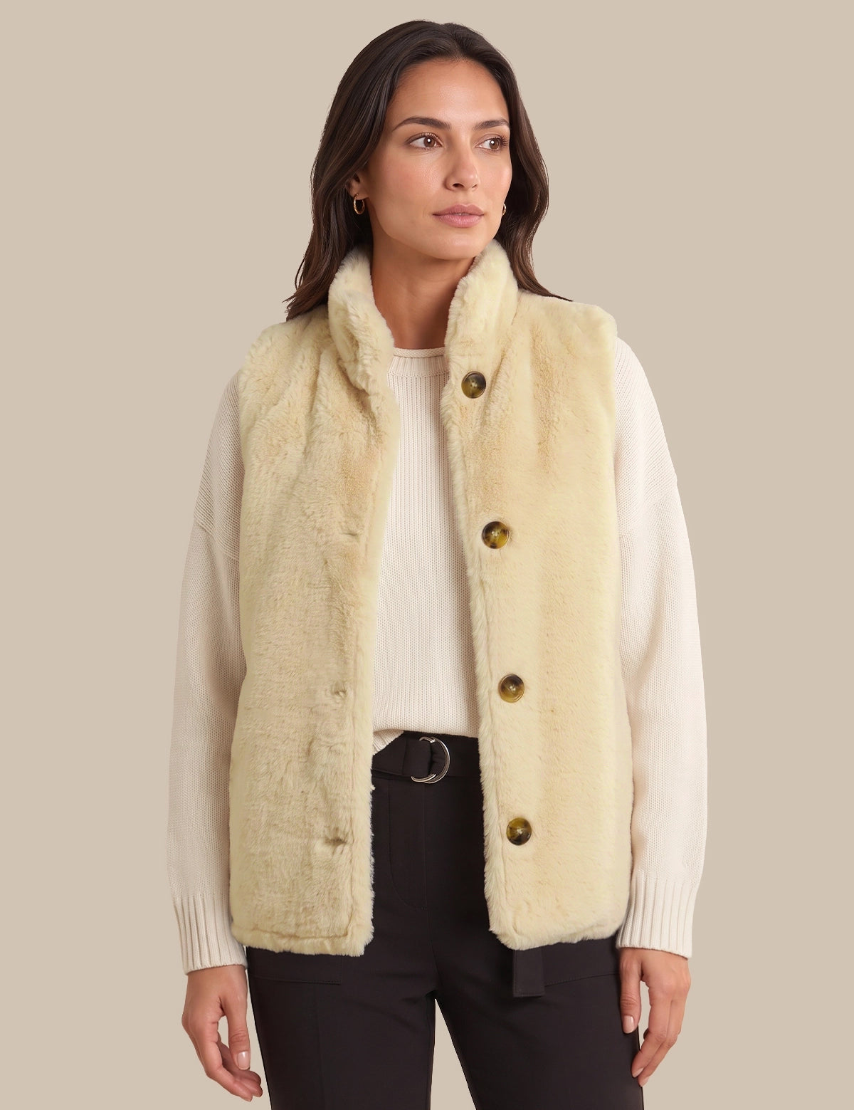 A woman modeling a cream-colored faux fur vest with large brown buttons over a white ribbed sweater and black pants, set against a solid beige background.
