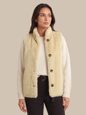 A woman modeling a cream-colored faux fur vest with large brown buttons over a white ribbed sweater and black pants, set against a solid beige background.