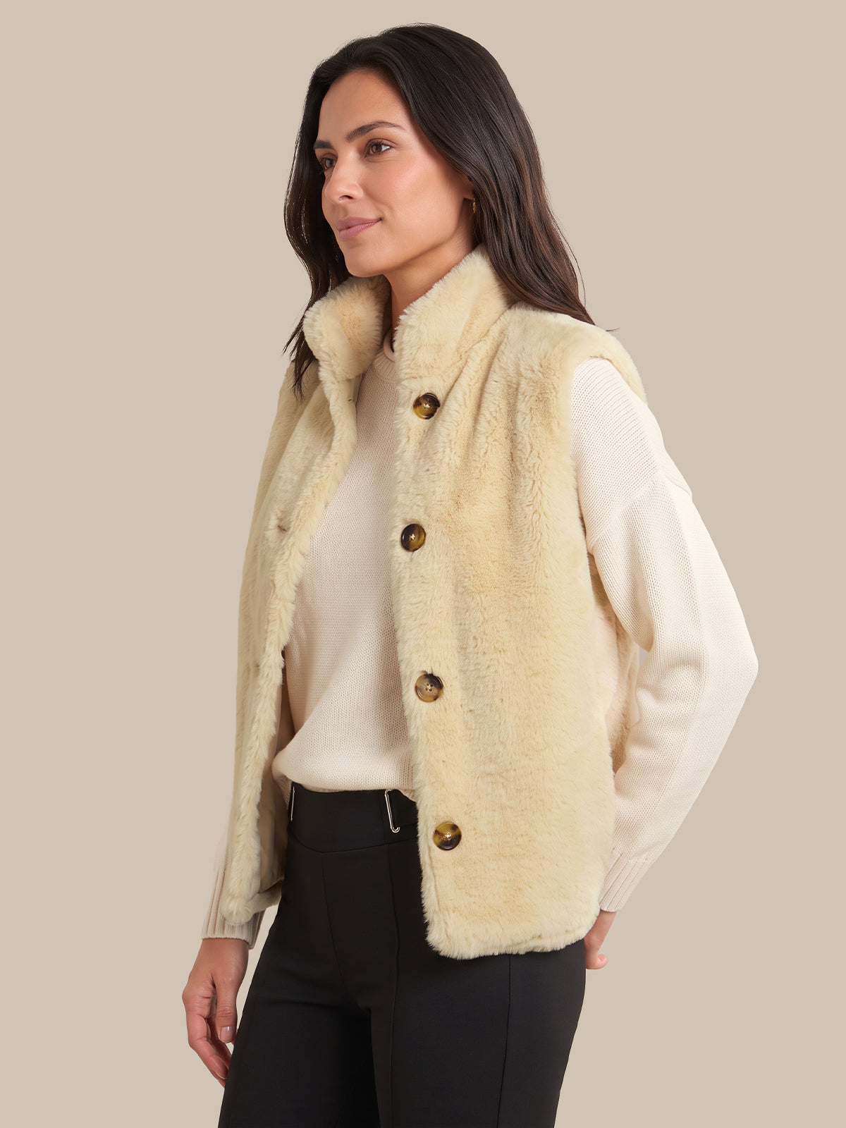A woman wearing a cream-colored faux fur vest with brown buttons over a cream sweater and black pants, photographed against a solid beige background.