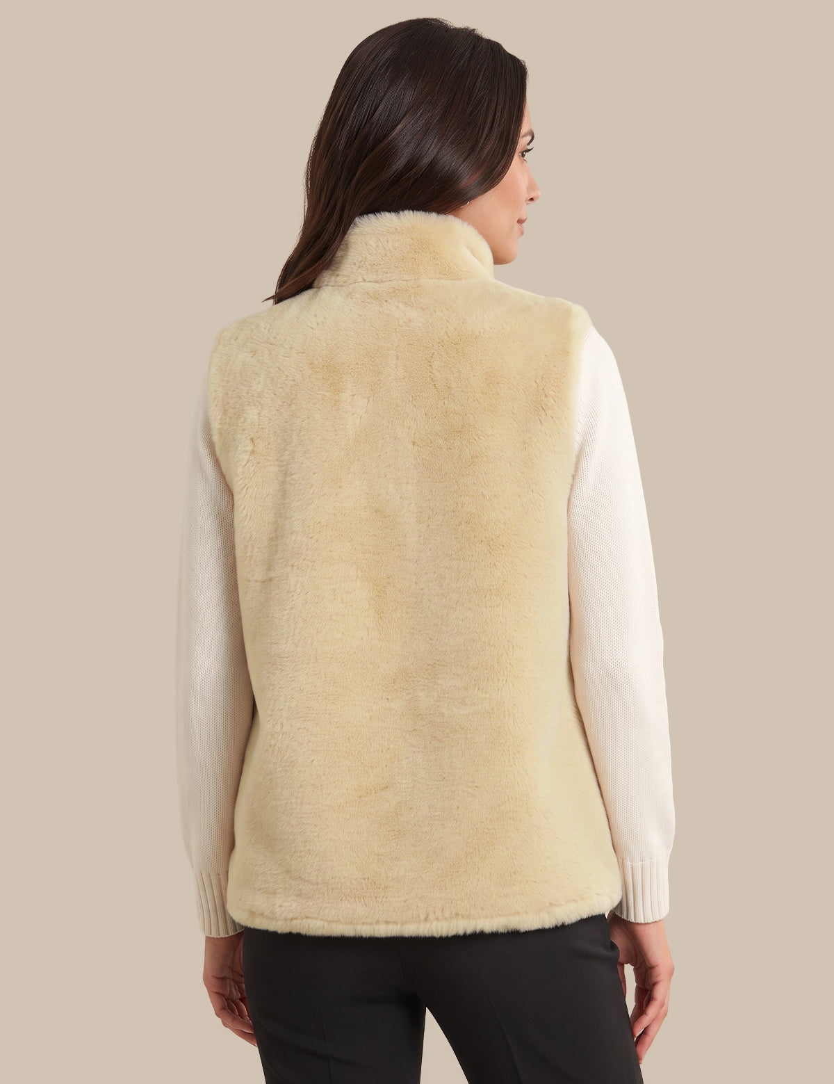 A woman shown from the back wearing a cream-colored faux fur vest over a white long-sleeve top and black pants, set against a solid beige background.