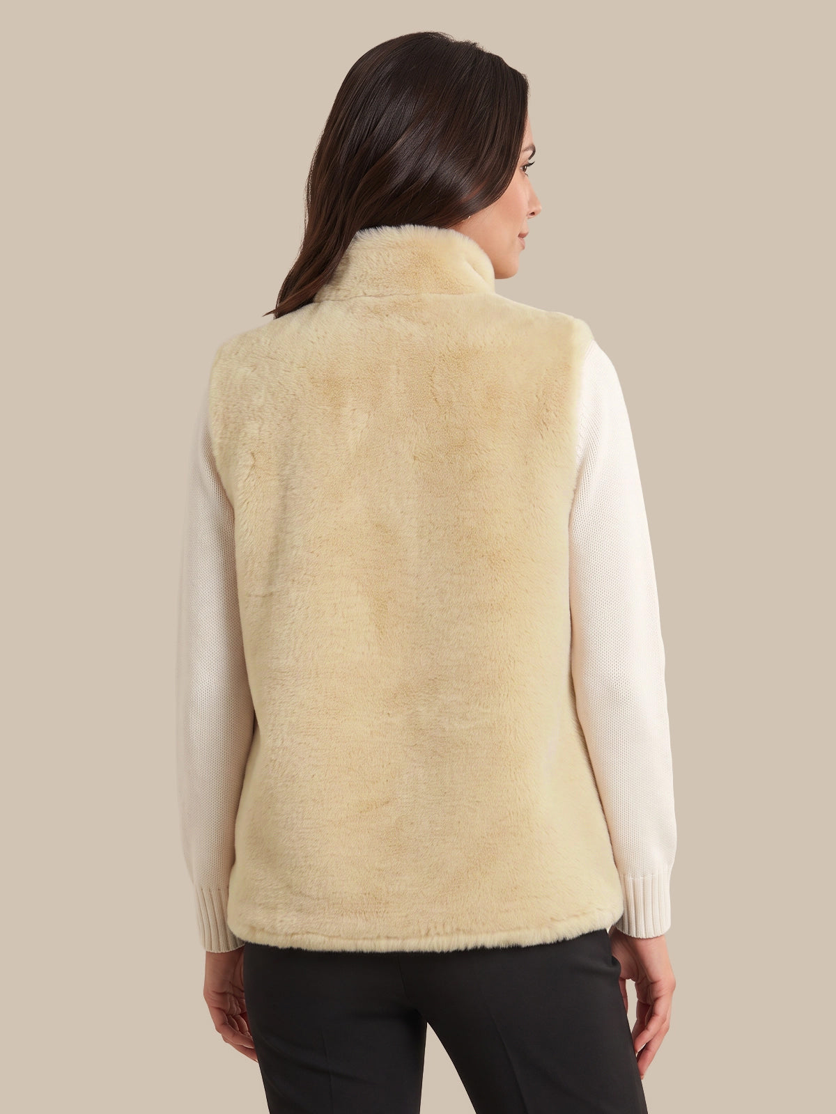 A woman shown from the back wearing a cream-colored faux fur vest over a white long-sleeve top and black pants, set against a solid beige background.