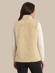 A woman shown from the back wearing a cream-colored faux fur vest over a white long-sleeve top and black pants, set against a solid beige background.