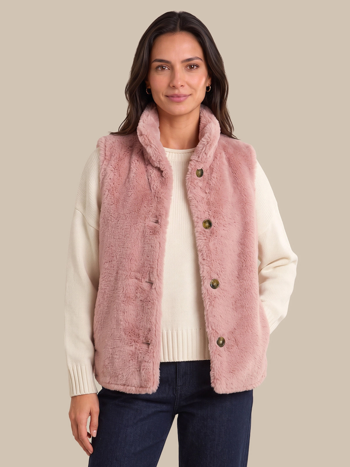 A woman modeling a dusty pink faux-fur vest over a cream ribbed sweater and dark blue jeans, set against a solid beige background.
