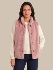 A woman modeling a dusty pink faux-fur vest over a cream ribbed sweater and dark blue jeans, set against a solid beige background.