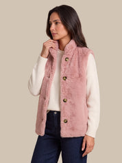 A woman wearing a dusty pink faux-fur vest over a cream ribbed sweater and dark blue jeans, posed against a solid beige background.