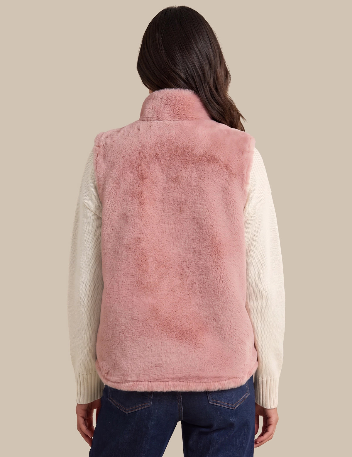 A woman shown from the back wearing a dusty pink faux fur vest over a cream-colored long-sleeve sweater and dark blue jeans, set against a solid beige background.