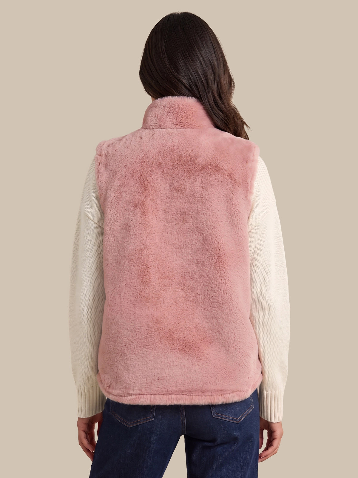 A woman shown from the back wearing a dusty pink faux fur vest over a cream-colored long-sleeve sweater and dark blue jeans, set against a solid beige background.