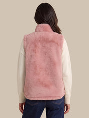 A woman shown from the back wearing a dusty pink faux fur vest over a cream-colored long-sleeve sweater and dark blue jeans, set against a solid beige background.