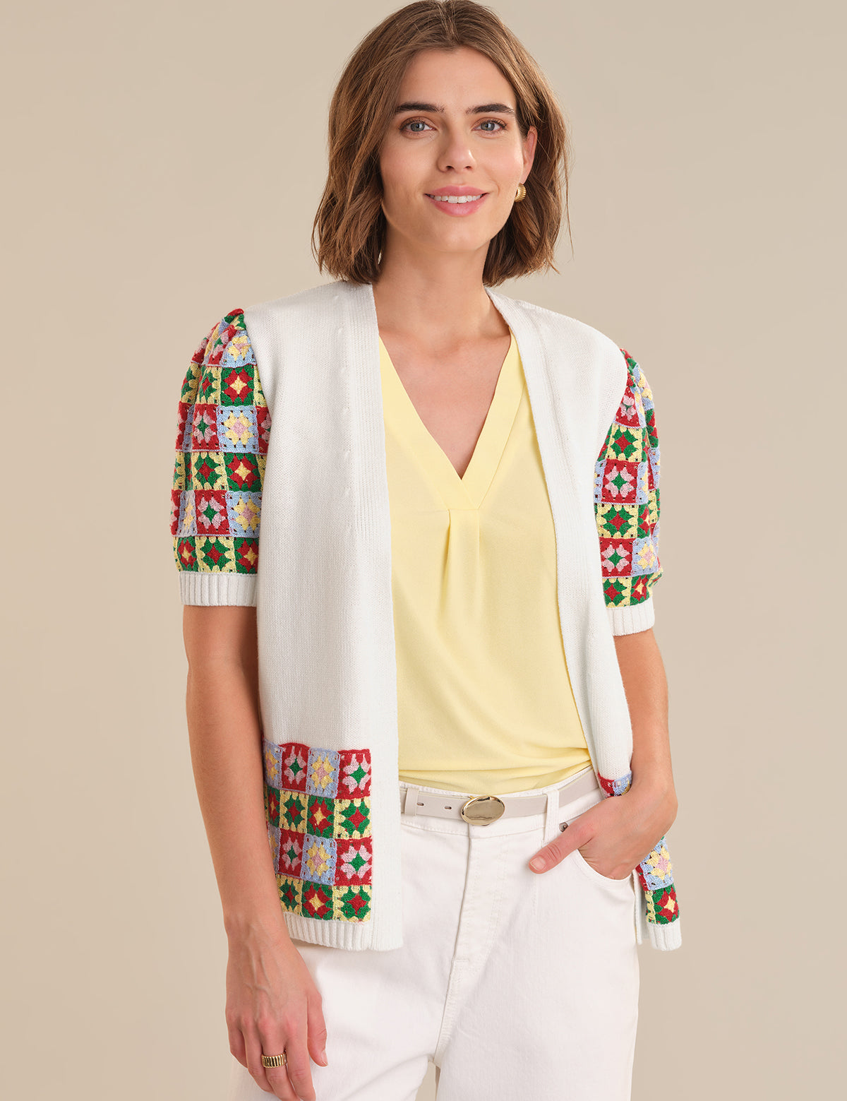 Woman wearing a colorful patterned vest over a yellow top and white pants on a beige background