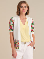 Woman wearing a colorful patterned vest over a yellow top and white pants on a beige background