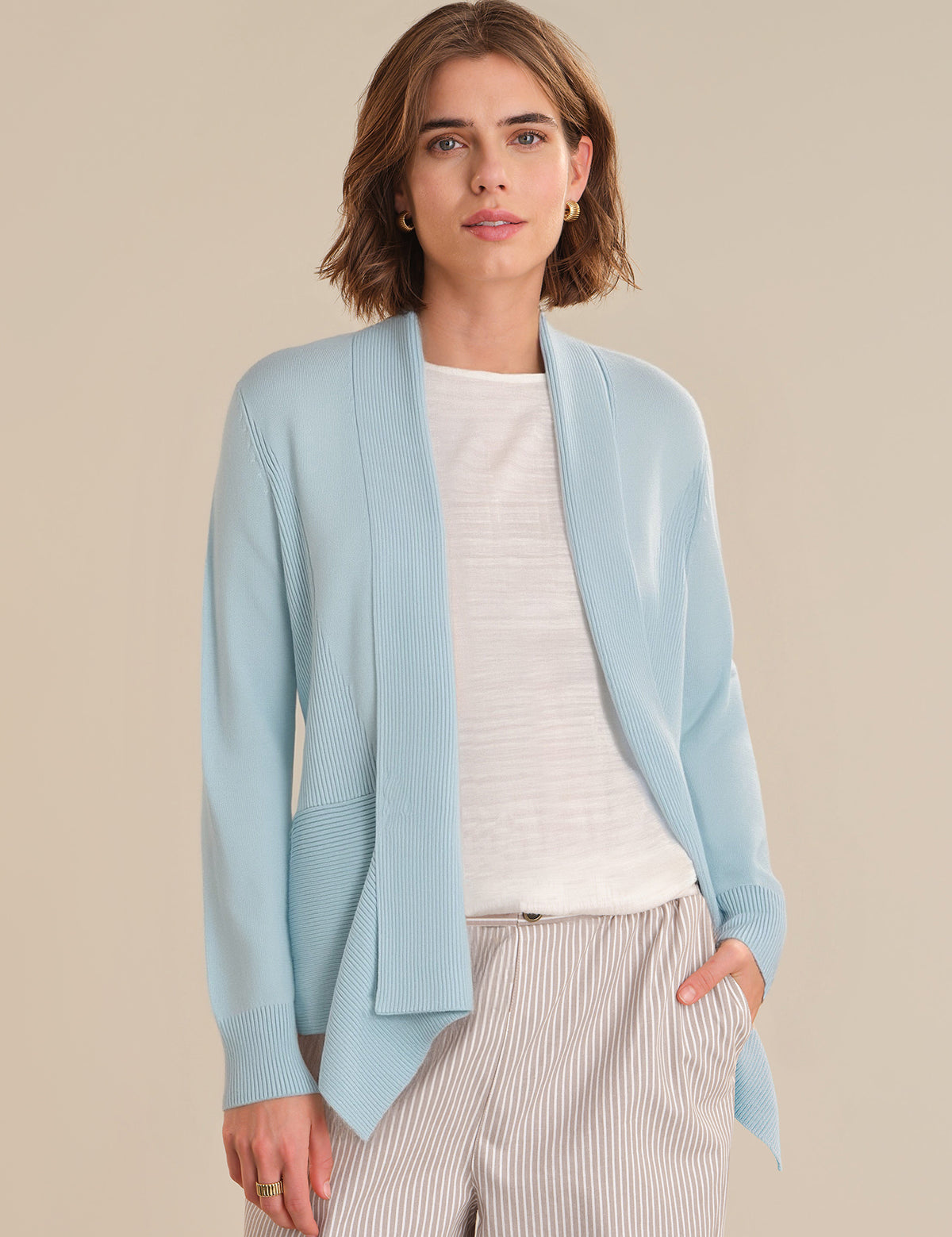 Woman wearing a light blue cardigan over a white shirt and beige pants on a beige background