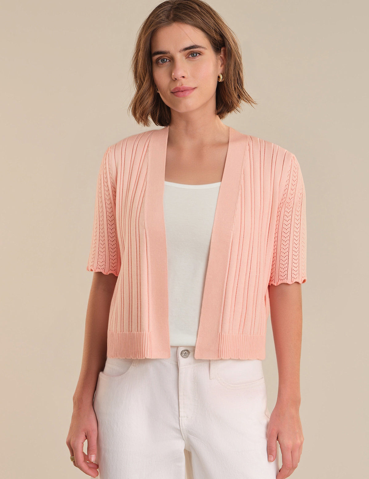 Woman wearing a light pink pleated shrug over a white top and pants on a beige background