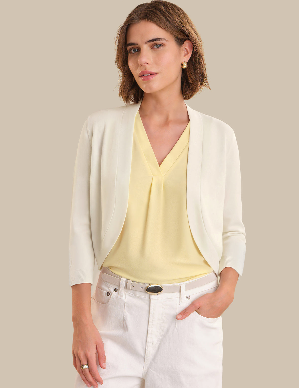 Woman wearing a white cardigan over a yellow top and white pants on a beige background
