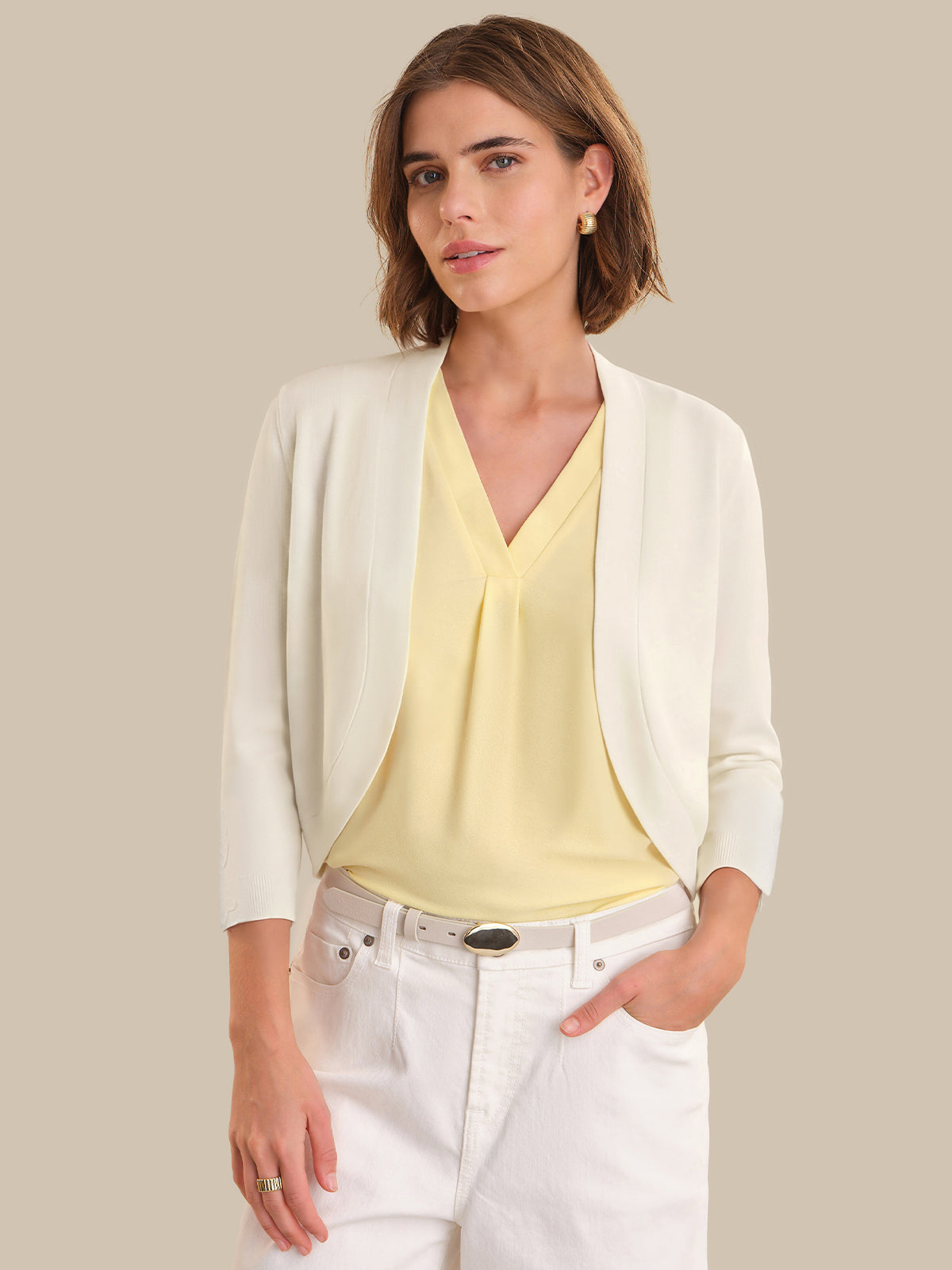 Woman wearing a white cardigan over a yellow top and white pants on a beige background