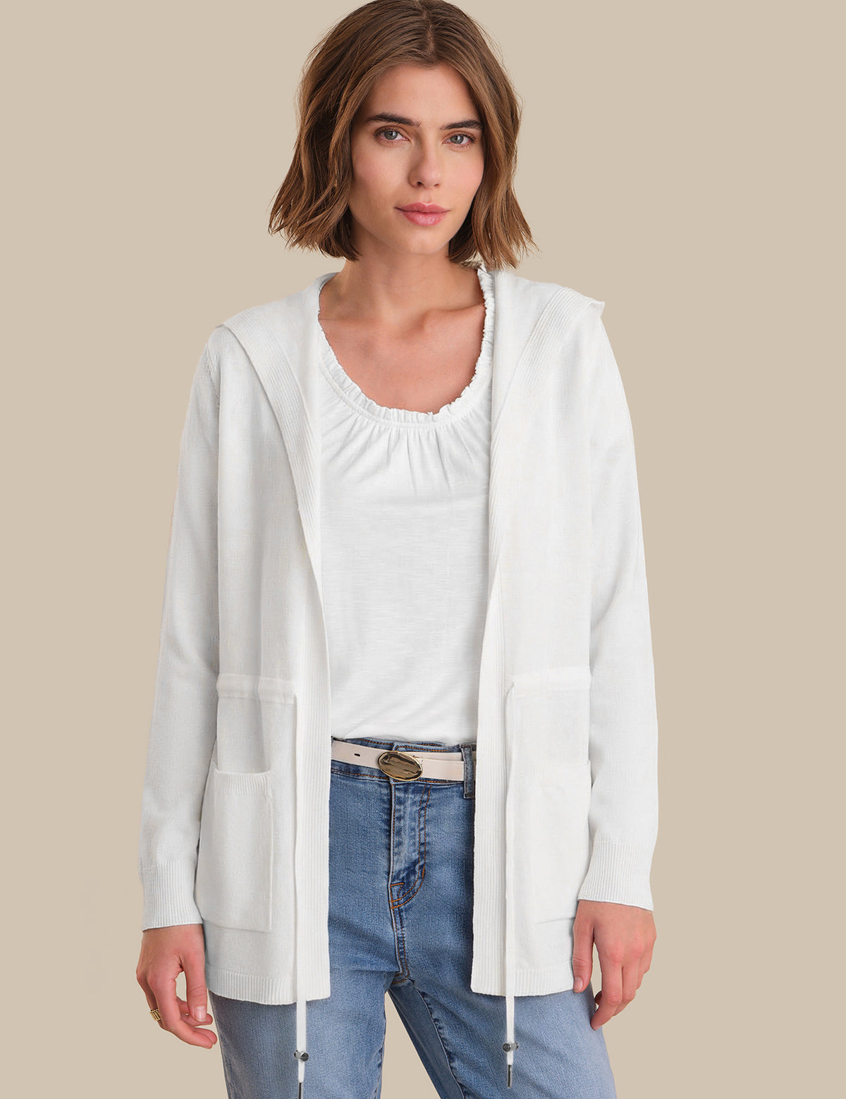 Woman wearing a white cardigan over a white top and blue jeans on a beige background