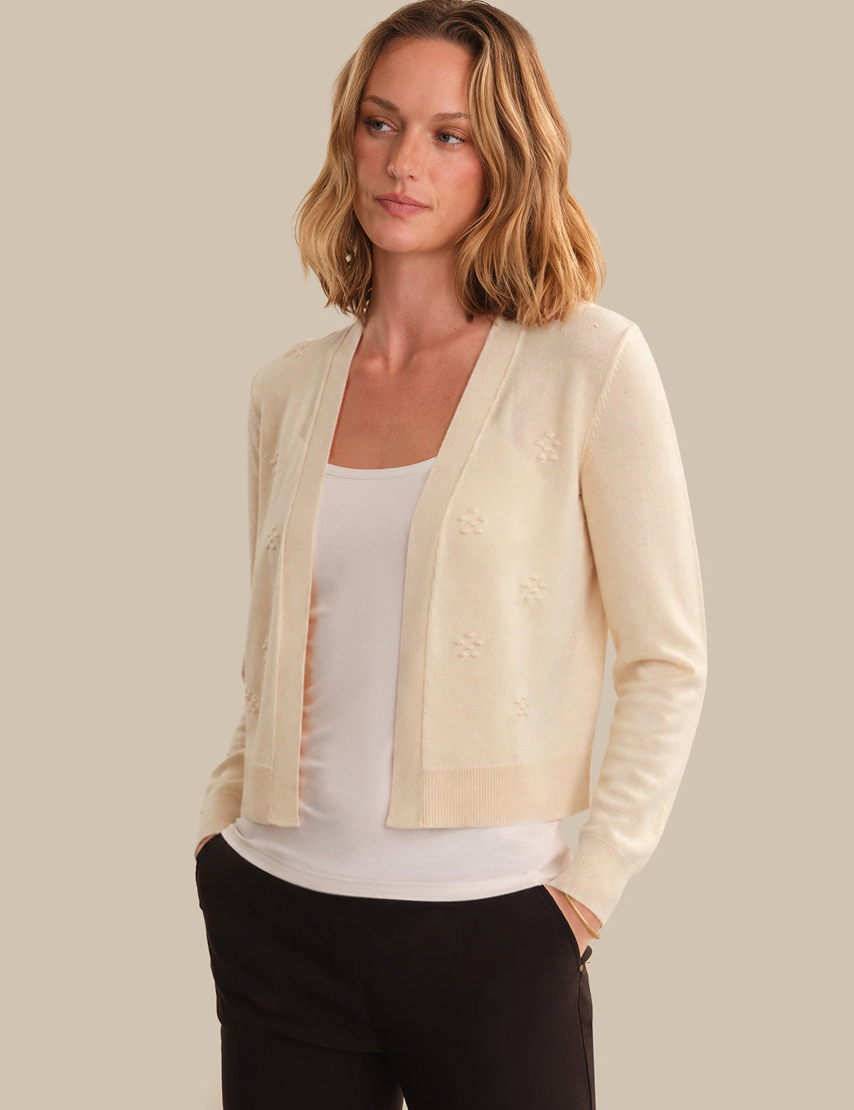 Woman wearing a light beige cardigan over a white top and black pants on a neutral background